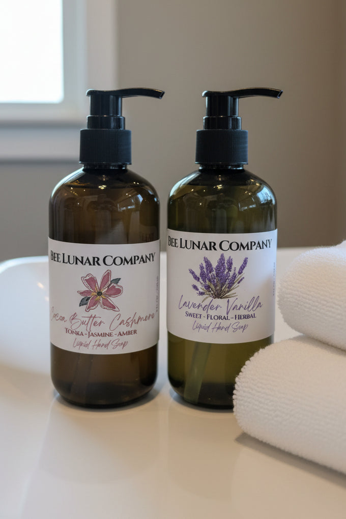 Two bottles of Bee Lunar Company liquid hand soap on a bathroom counter with towels in the background.