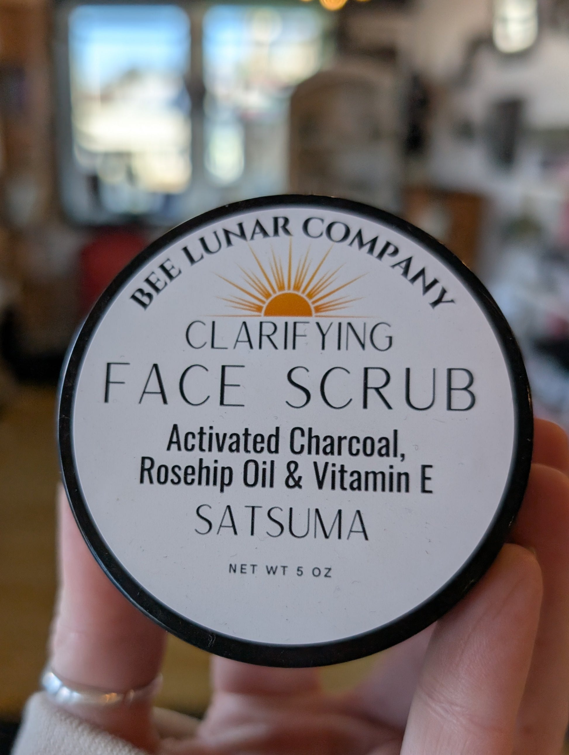 Hand holding a container of Bee Lunar Company Clarifying Face Scrub with a blurred indoor background.