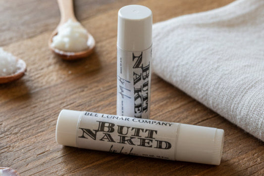 Two lip balm tubes labeled 'Butt Naked' and 'Bee Lunar Company' held by a hand in a blurred indoor setting.